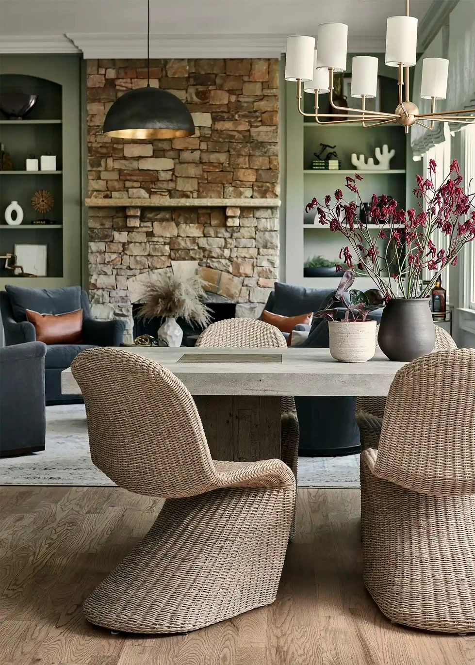 keeping room | sitting room | living room | family room | blue upholstered swivel armchair | blue arm chair | round wood coffee table | brass bar cart | metal dome pendant light | black and brass dome pendant light | green builtin bookcase | stacked stone fireplace | round black end table | round black side table | wicker chair | dining table and chairs | dining chairs | wicker dining chairs | concrete table top | stone table top | kitchen table