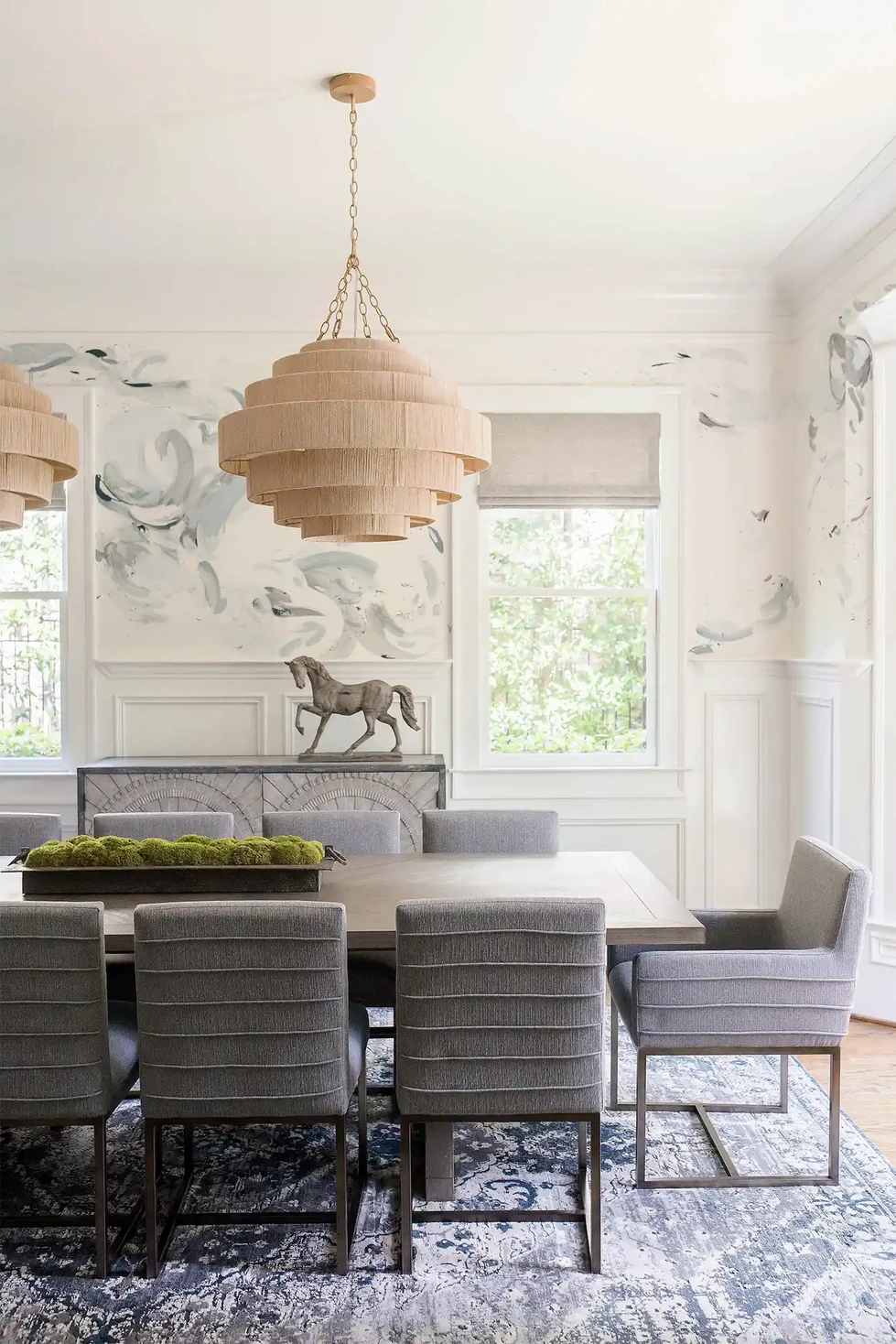 dining room interior design | dining table and chairs | hand painted wall mural | custom abstract wall painting | gray upholstered chairs | blue area rug | gray stone credenza buffet sideboard | oversized large wood dining room table | Palecek Everly woven rope pendant | dark stained wood floor
