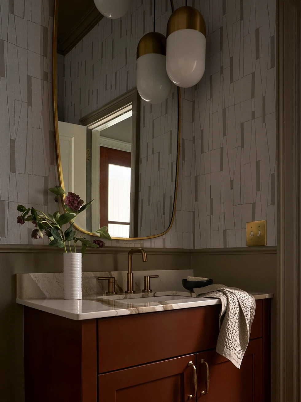 Bathroom vanity with warm wood cabinet, marble countertop, brass faucet, rounded mirror, and soft wall sconces against patterned wallpaper