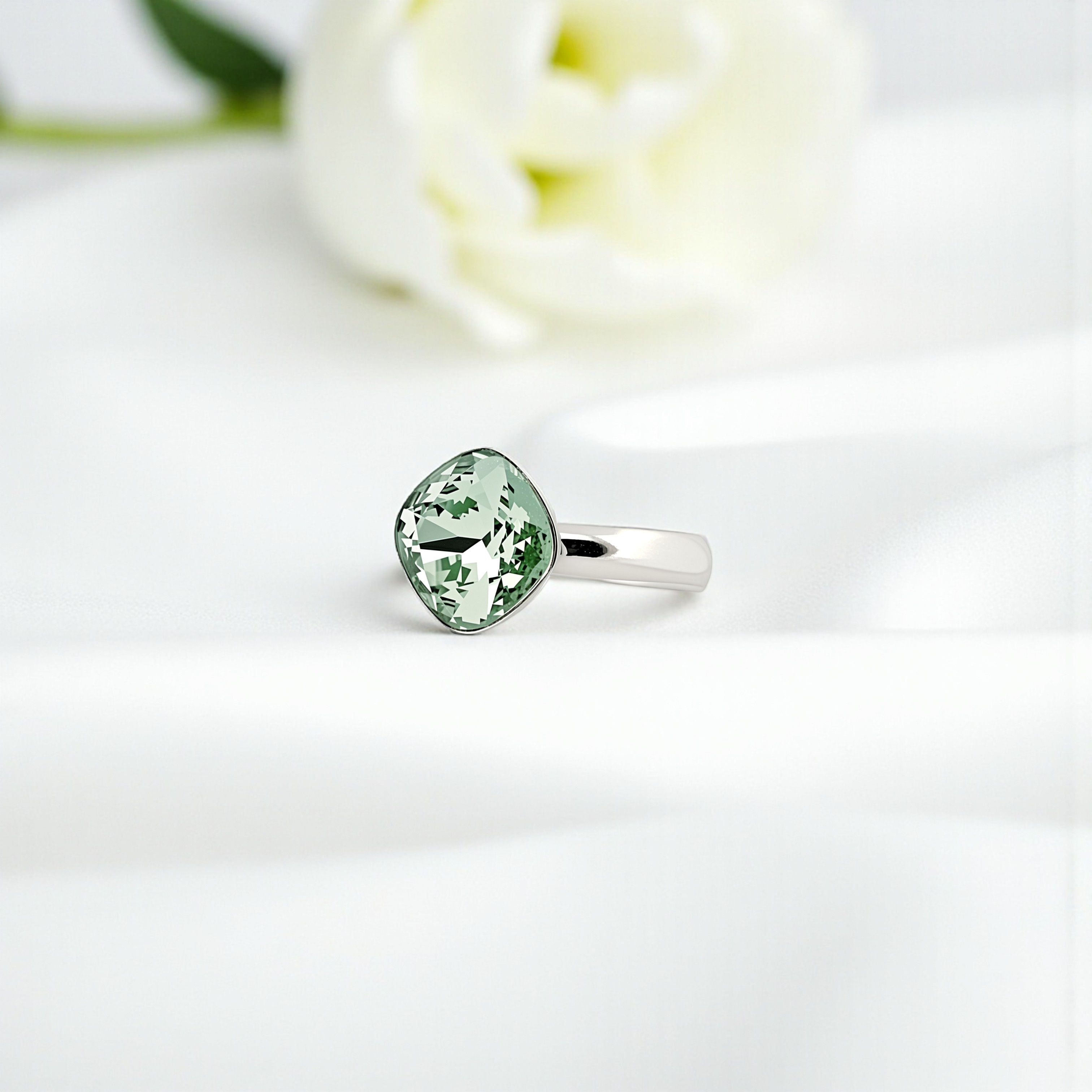 Bague en argent 925 ornée d’un cristal Swarovski® vert Chrysolite de 12 mm, posée sur tissu blanc avec fleur blanche.