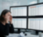 Woman stressed at desk, surrounded by four monitors displaying spreadsheets. Crumpled paper and coffee cup visible. Whiteboard text in background.