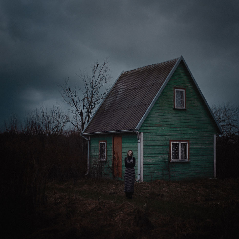 A gothic-inspired photograph of a person in dark clothing standing before a lonely green wooden house in a desolate, moody landscape.