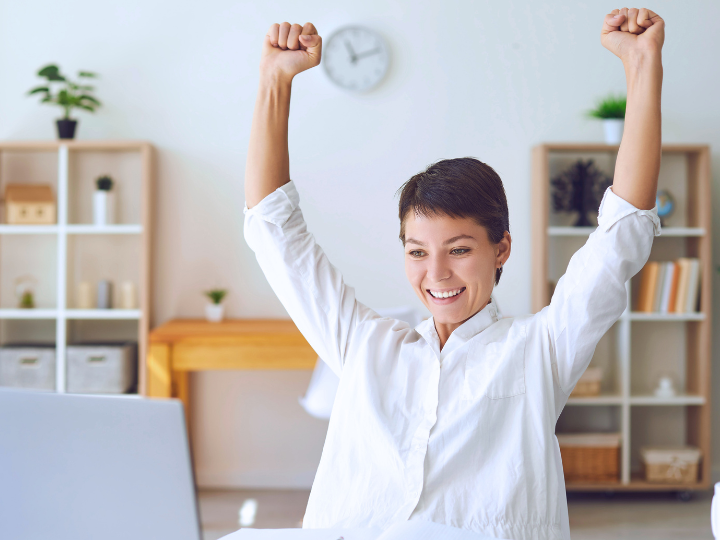 Smiling female entrepreneur in white shirt with raised fists celebrates at a desk. Shelves and plants in bright room, clock on wall.