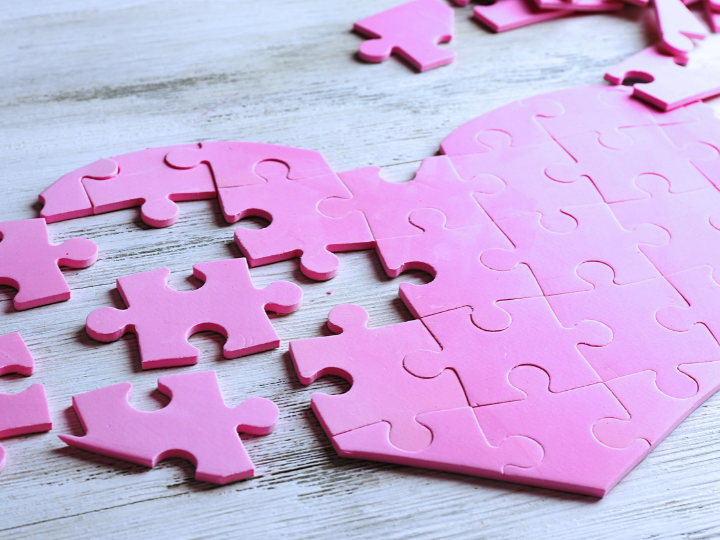 Pink jigsaw puzzle pieces arranged into a heart on a white wooden surface, with a few pieces still scattered nearby.