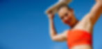 Personal trainer wearing orange sports bra smiling and holding a towel over her head with blue sky in the background.