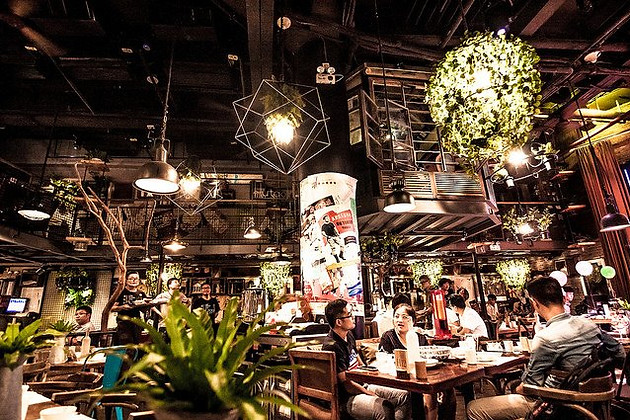 Z Square Group. The image depicts a busy restaurant with a large dining area filled with tables and chairs. There are several people seated at the tables, enjoying their meals. The walls are adorned with hanging plants, adding a touch of greenery to the space. The lighting in the restaurant is dim, creating a cozy and inviting atmosphere.