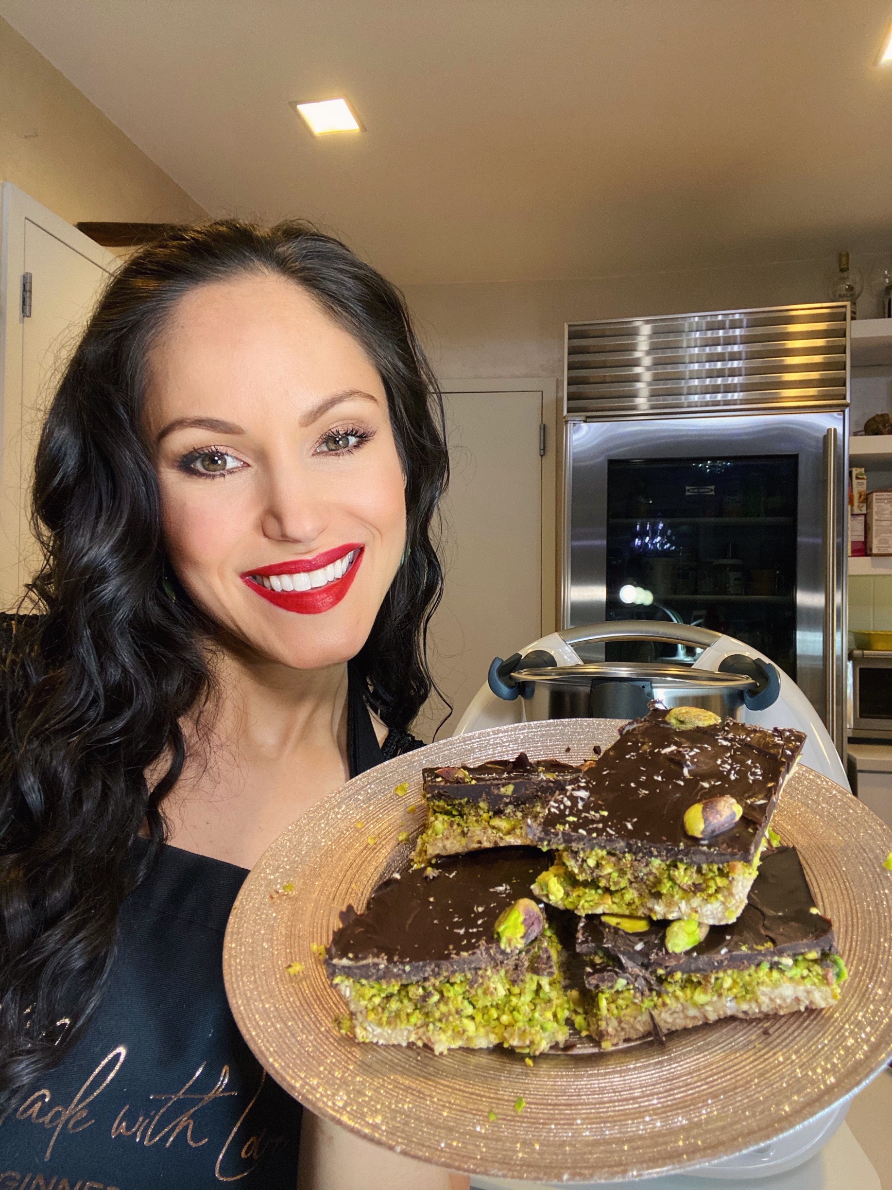 vegan chef cheryl martinez holds a plate of chocolate and pistachio bars in a kitchen. She's wearing a black apron made with love. Warm lighting creates a cozy mood.
