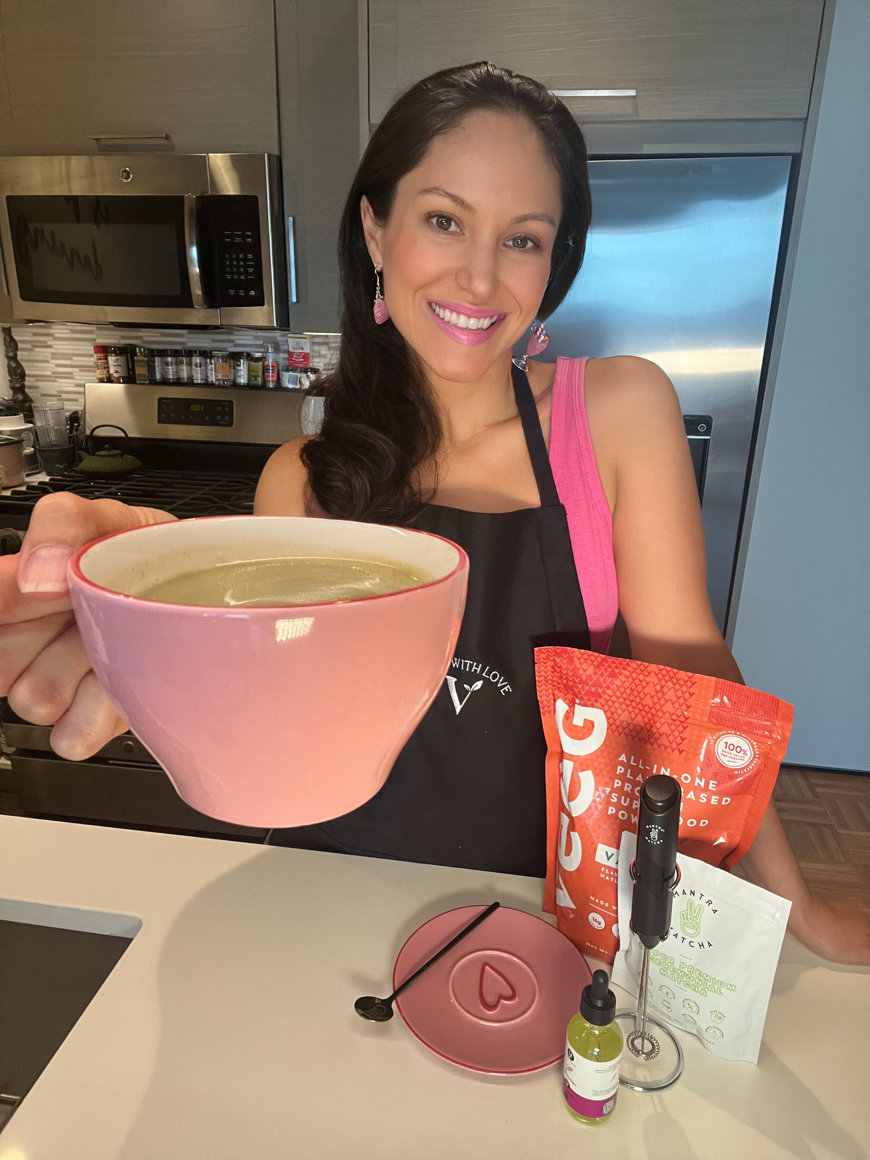 Woman smiling in kitchen holding pink bowl, wearing apron. Counter has vegan powder, tea packet, dropper bottle.