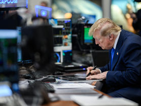 President Donald Trump leans over a desk at NASA Mission Control Center in Houston, writing on a document with computer monitors around him.