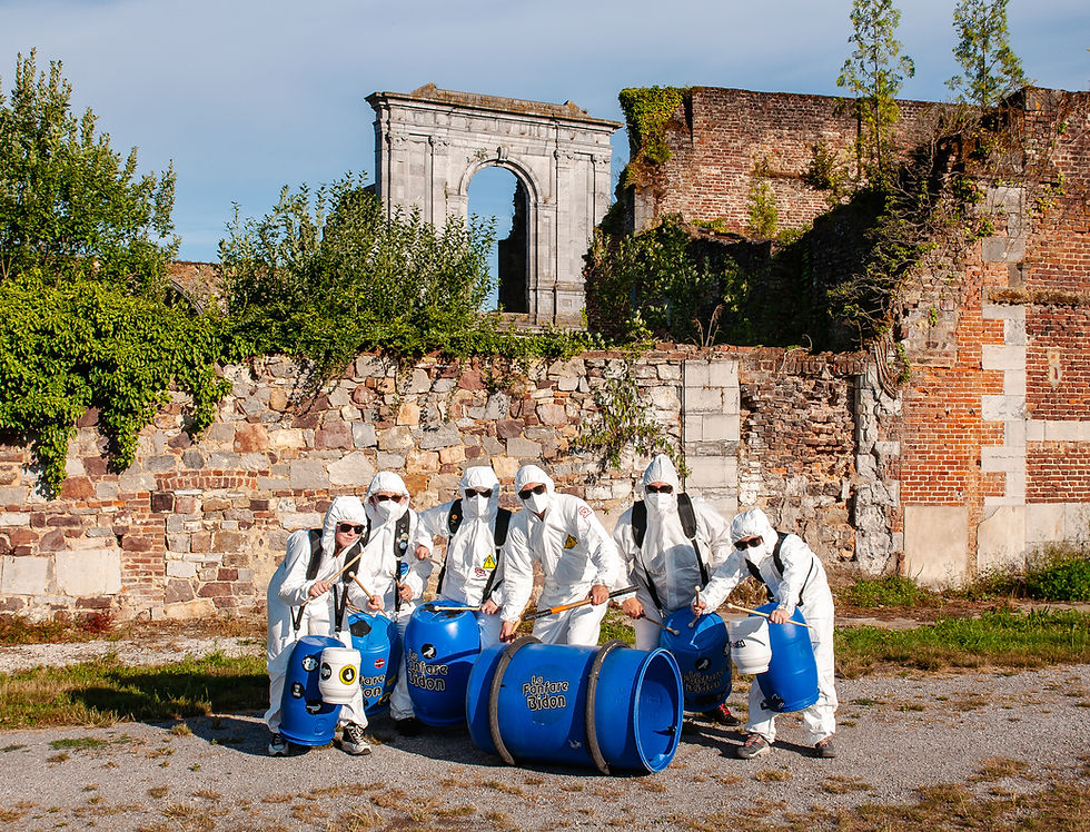 Miniature : Équipe en combinaisons protectrices devant des barils bleus et un bâtiment en ruine.