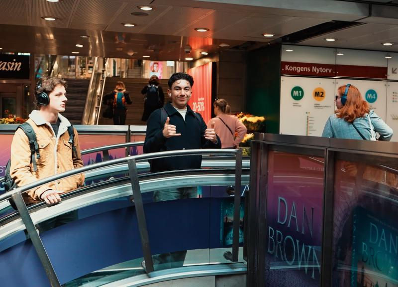 Two people with headphones in a subway station. One gives thumbs up; the other looks aside. Signs read "Kongens Nytorv" and "Dan Brown."