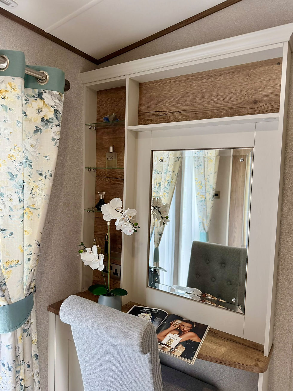 ABI Beaumont bedroom dressing area with a mirror, open shelving, an upholstered chair and floral curtains.