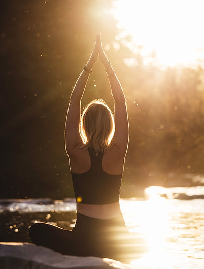 Karin in der Meditationshaltung von hinten Fotografiert. Am Fluss mit dem Sonnenuntergang im Hintergrund.