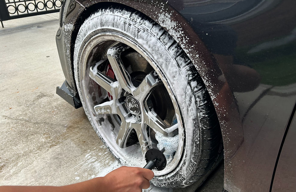 Hand scrubs a soapy alloy car wheel with a brush on a driveway.