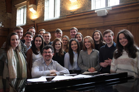 Group photo of Choral Scholars and Desmond Earley standing around a piano