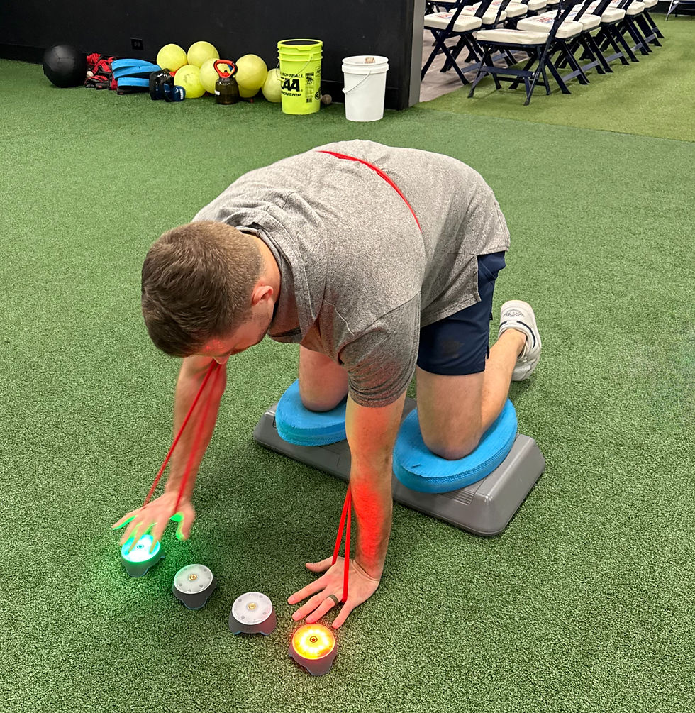 Athlete kneels on turf using glowing training lights with resistance bands in an indoor gym, with folding chairs and exercise gear behind.