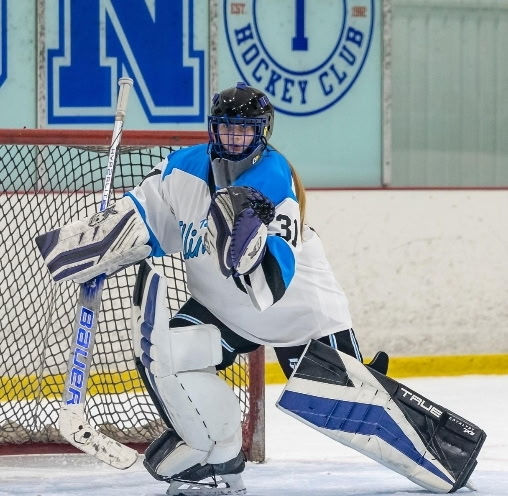 Female hockey goalie in white and blue gear crouches in net on ice rink, with N Hockey Club sign behind.