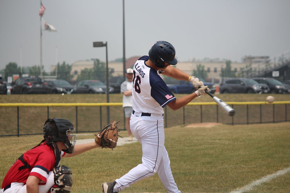 Baseball batter in white swings at a pitch as catcher in red crouches behind, on a grassy field with parked cars and flags.