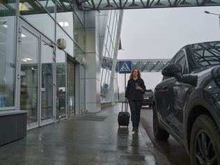 A woman in dark clothing walks with a suitcase outside a glass airport terminal. Overcast sky and cars in the background create a gloomy mood.