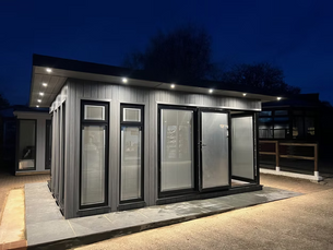 Modern gray modular building with black trim and glass doors glowing under roof lights at dusk.
