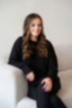 Smiling woman with long, wavy hair sits on a light couch wearing a black top. She has pink nails and rings, exuding a relaxed mood.