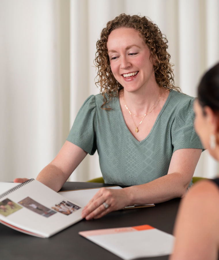 A speech-language pathologist smiles across a table at her client during a language therapy session, an open workbook in front of her.
