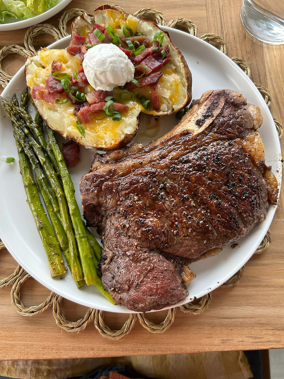 Plate with a juicy steak, grilled asparagus, and a loaded baked potato topped with sour cream, bacon, and chives on a wooden table.