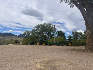 Two green tractors and farm equipment parked on a gravel ranch road under a cloudy sky, with trees and mountains in the background.