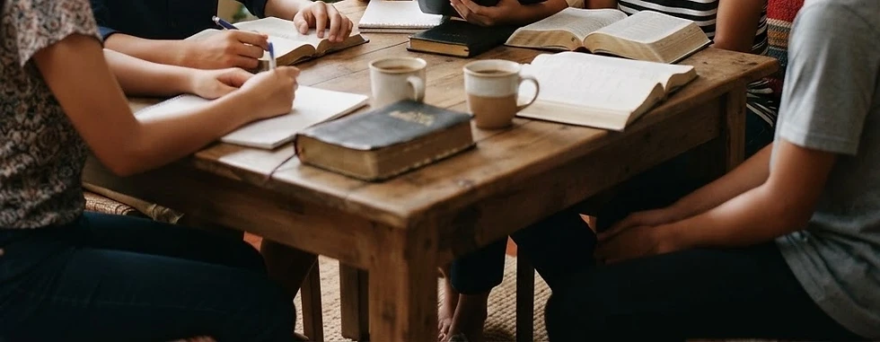 Lifestyle photography of a diverse group of young adults studying around a wooden table in a home setting. The atmosphere is peaceful and scholarly, with books and open Bibles spread across the table.