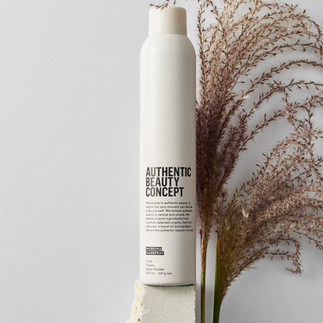 Eine beige Haarsray-Dose mit schwarzer Aufschrift steht auf einem Stein vor einem hellgrauen Hintergrund. Auch ein Trockenblumen-Strauch steht im Hintergrund.