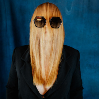Auf dem Foto sieht man eine Frau in schwarz gekleidet. Ihre rotblonden Haare sind nach vorne über das Gesicht gekämmt. Sie trägt eine große schwarze Brille. Der Hintergrund ist petrolgrün.