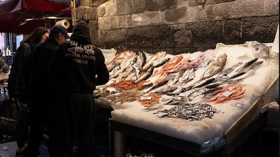 Marktszene mit Menschen, die frischen Fisch an einem Stand unter rotem Zelt begutachten. Hintergrund: alte Steinmauer. Lebhafte Stimmung.