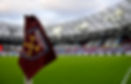 Football stadium with a burgundy West Ham United corner flag in the foreground, green field, and empty stands under a cloudy sky.