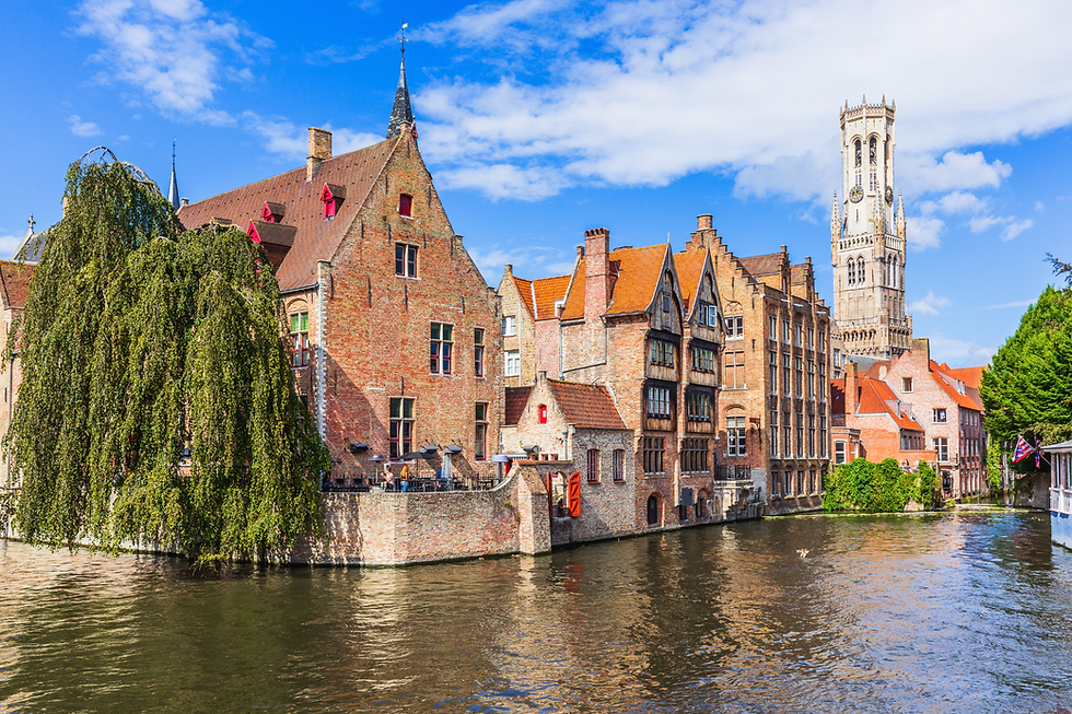 Sunny canal with medieval brick houses and a tall church tower in Bruges under a blue sky, calm water reflecting buildings.