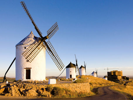 White windmills on a golden hill under a clear blue sky, with a winding road and a distant stone fortress at sunset.