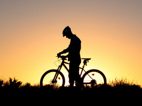 Silhouette of a person with a bike at sunset. They stand on grass, sky glowing with orange and purple hues, creating a calm mood.