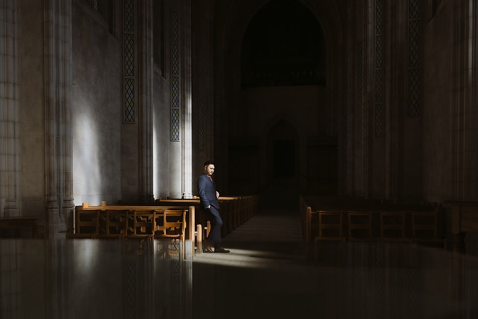 Groom leaning against a wooden pew in a shaft of window light at Trinity College Chapel in Toronto.