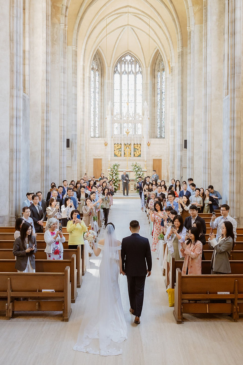 Bride and groom seen from behind, holding hands in the Trinity College Chapel aisle with guests on both sides.