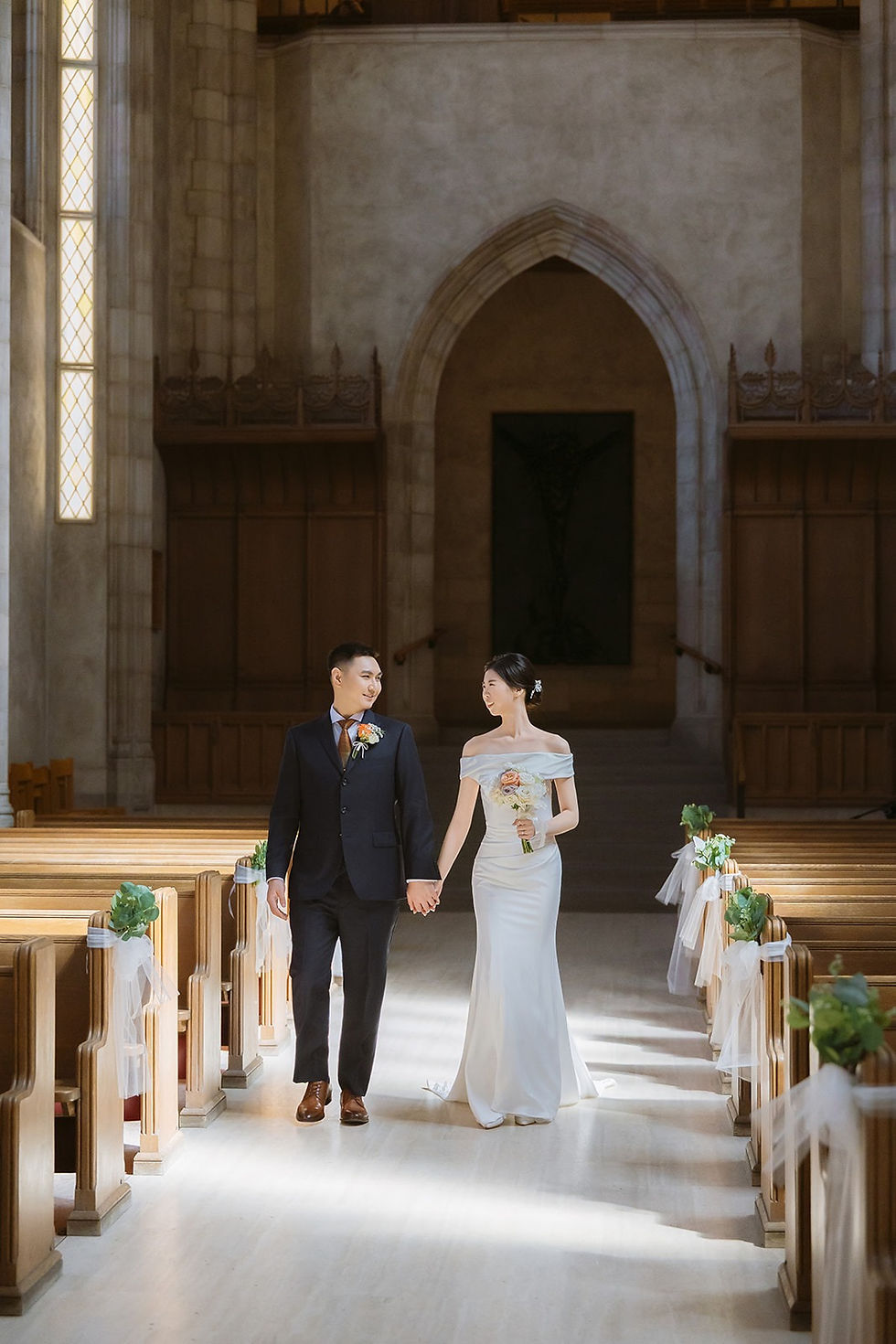 Couple smiling at each other as they walk hand in hand through sunlight in the Trinity College Chapel aisle.