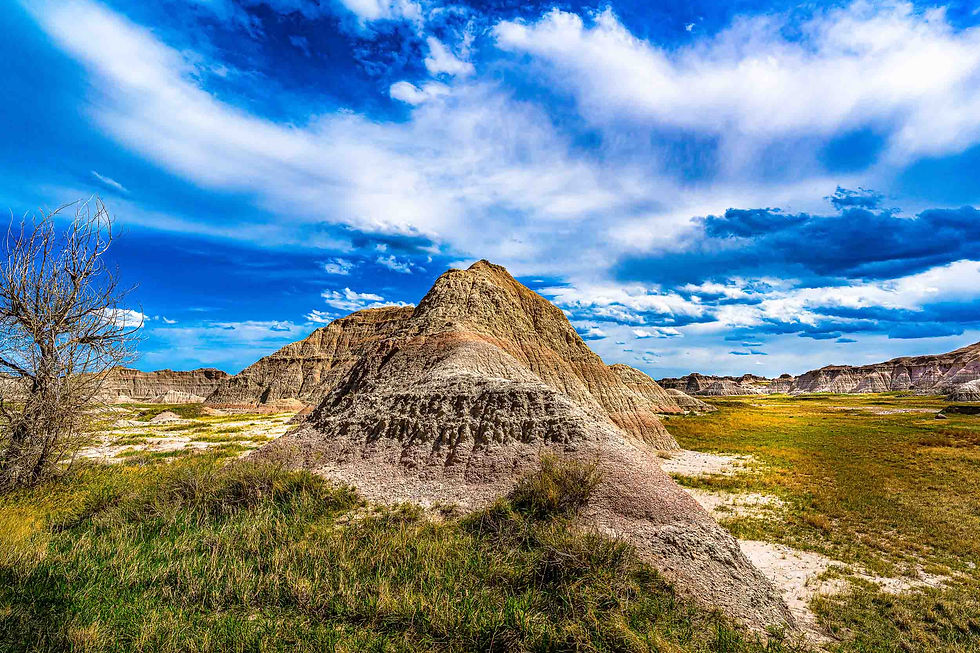 A small mountain in Badlands National Park with surrounding rock formations and green meadows of grass with a cloudy sky.