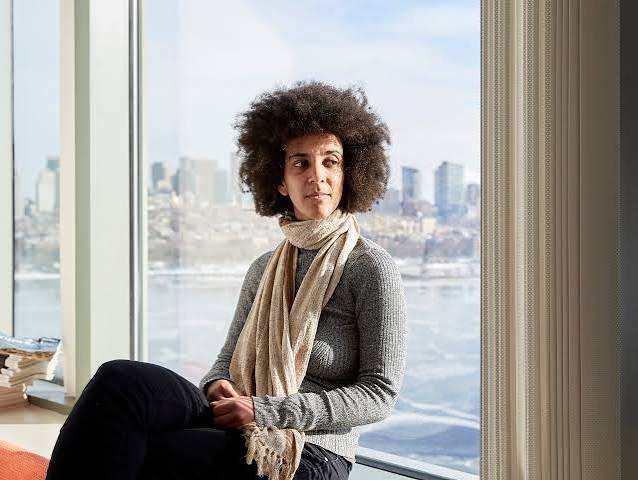 Color landscape photo of Timnit Gebru sitting by a large window with a city skyline and river in the background
