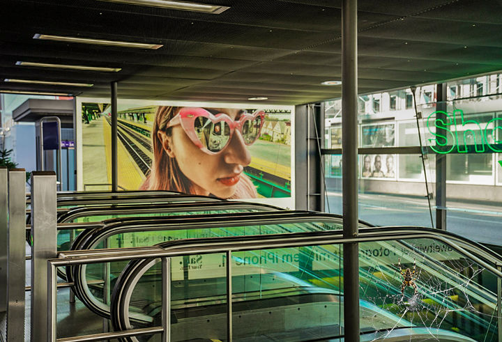 Fotocomposing mit Rolltreppen, einem grossen Werbeplakat mit Frauenportrait und einer Spinne im Netz im Vordergrund.