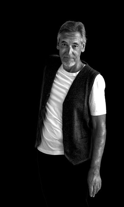 Schwarzweiss-Portrait eines Mannes mit weissem T-Shirt und schwarzer Weste vor schwarzem Hintergrund