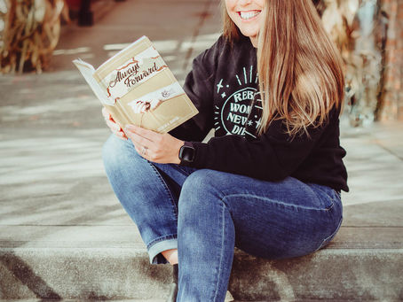 Female with light brown hair, sitting on steps, holding an open book and smiling.