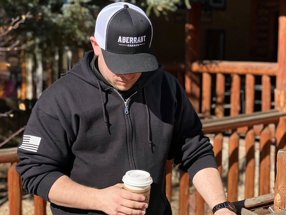Man holding a paper coffee cup standing in front of a wooden cabin's banister, wearing a black and white Aberrant Garage hat and a zip up hoodie with an American Flag on the left sleeve.