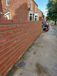 Mason constructing a new red brick wall along a paved walkway.