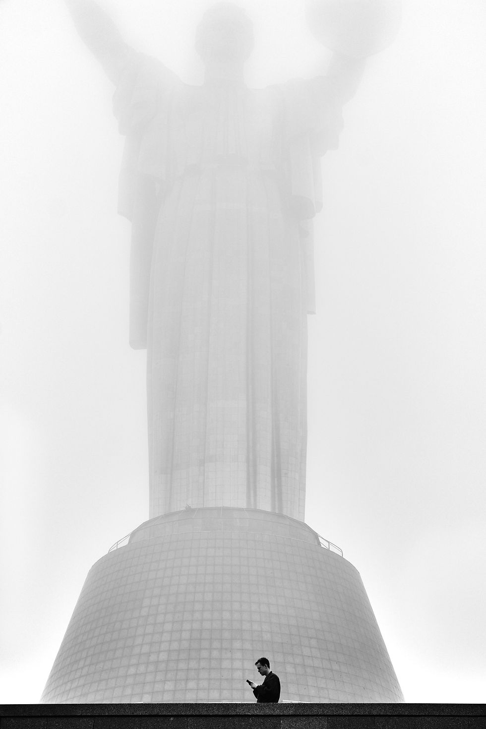 Muž stojí s telefonem v ruce před monumentální sochou Matka Ukrajina v Kyjevě