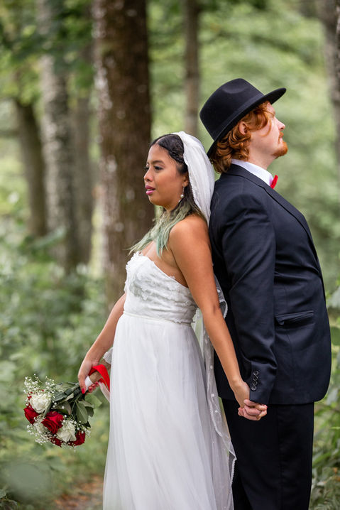 Utomhusbröllop där bröllopsparet står i skogen. Dem håller varandra i handen med ryggarna mot varandra. Fotat av Oskar Gillblad i Göteborg. 