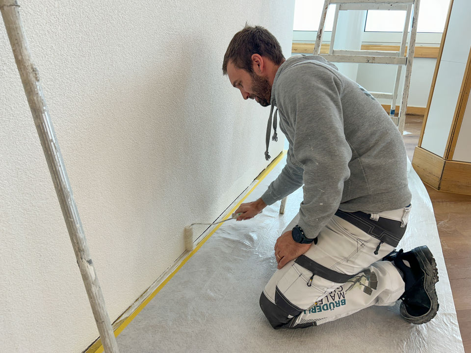Maler kniet am Boden und klebt eine Wand mit Malerklebeband ab.