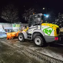 Radlader räumt Schnee, NEXUS, grünes Logo mit orangefarbenem Schneepflug nachts.